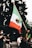 A person in a crowd raises a large Mexican flag with a green, white, and red color scheme, featuring an eagle emblem in the center. The scene shows multiple people, some wearing masks and gloves, possibly indicating a protest or public gathering. The background includes trees and the base of a lamp post.