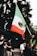 A person in a crowd raises a large Mexican flag with a green, white, and red color scheme, featuring an eagle emblem in the center. The scene shows multiple people, some wearing masks and gloves, possibly indicating a protest or public gathering. The background includes trees and the base of a lamp post.
