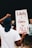 A person stands with their back to the camera holding a sign that reads 'Liberty & Justice for ALL!' with bold, red lettering. The individual is wearing a white t-shirt and a blue headwrap. The background shows an out-of-focus group of people, indicating a probable protest or gathering.
