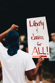 A person stands with their back to the camera holding a sign that reads 'Liberty & Justice for ALL!' with bold, red lettering. The individual is wearing a white t-shirt and a blue headwrap. The background shows an out-of-focus group of people, indicating a probable protest or gathering.