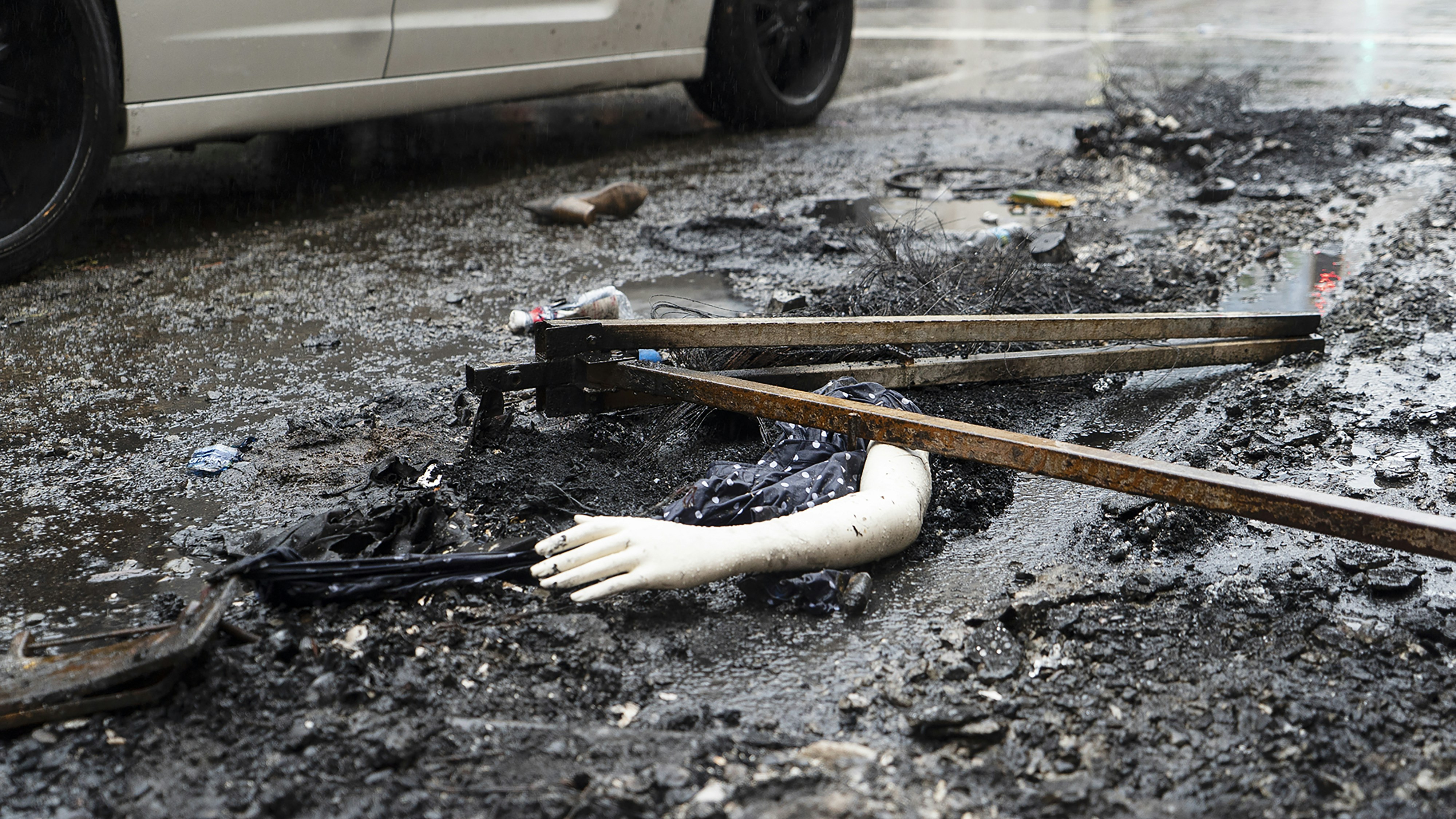 A close-up shot of a police officer collecting evidence from the ground near the burnt vehicle, wearing gloves.