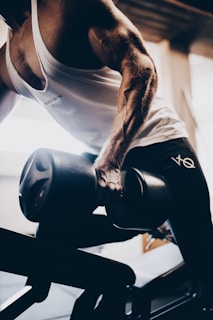 A muscular person in a tank top is working out with a dumbbell on a bench, focusing on their arm muscles. The lighting creates strong contrasts and highlights the definition of the musculature.