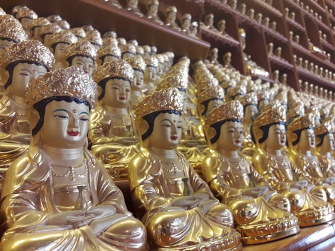 A multitude of identical golden Buddha statues are neatly arranged on shelves, each displaying intricate detailing, serene facial expressions, and sitting in a meditative posture. The statues are adorned with ornate headpieces and feature rich golden hues that create an aesthetically harmonious and spiritually calming atmosphere.