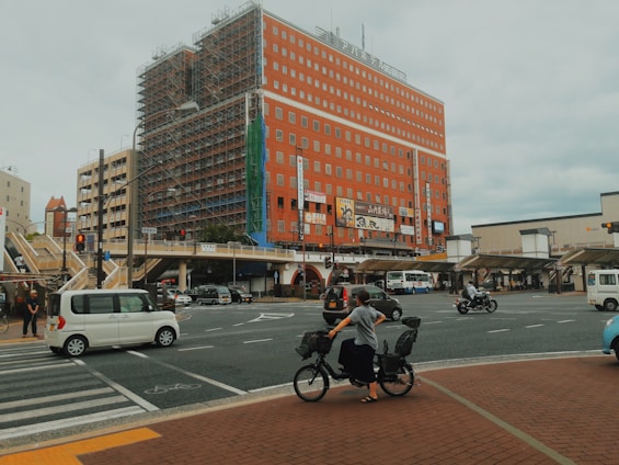 An urban scene with a large red-brick building under construction, featuring scaffolding and billboards. A busy intersection is visible, with cars, a person on a bicycle, a motorbike, and some pedestrians. The sky is overcast, and the streets appear active with various everyday activities.