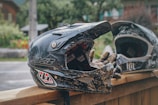 A close-up of two black motocross helmets resting on a wooden surface with gloves placed inside one. The setting appears to be outdoors with greenery and a building visible in the blurred background.