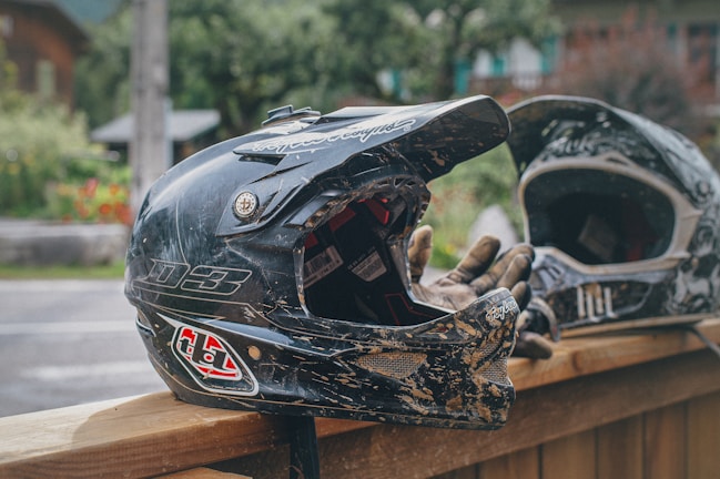 A close-up of two black motocross helmets resting on a wooden surface with gloves placed inside one. The setting appears to be outdoors with greenery and a building visible in the blurred background.