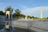 A large outdoor memorial with a prominent obelisk standing tall in the background. The foreground features a fountain surrounded by people walking and relaxing. Stone architecture and green trees complement the scene, and an American flag is visible prominently.