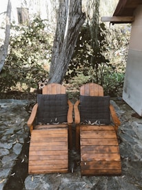 Two wooden lounge chairs with dark cushions are placed side by side on a stone patio. The chairs are situated next to a large tree and a shrub, creating a serene outdoor setting. Fallen leaves are scattered on the ground and on the chairs, adding to the rustic and natural ambiance.