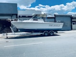 A white motorboat named 'Top Notch' is parked on a trailer in an industrial area. The trailer is positioned on a street in front of a building with metal shutters. The sky is clear with a few clouds.