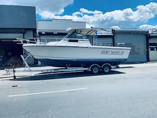 A white motorboat named 'Top Notch' is parked on a trailer in an industrial area. The trailer is positioned on a street in front of a building with metal shutters. The sky is clear with a few clouds.
