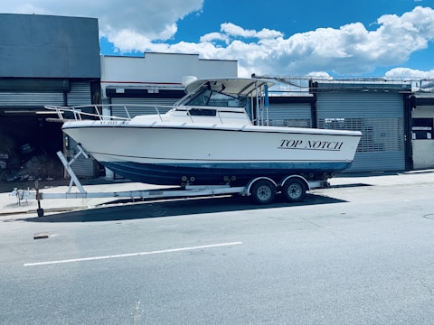 A white motorboat named 'Top Notch' is parked on a trailer in an industrial area. The trailer is positioned on a street in front of a building with metal shutters. The sky is clear with a few clouds.