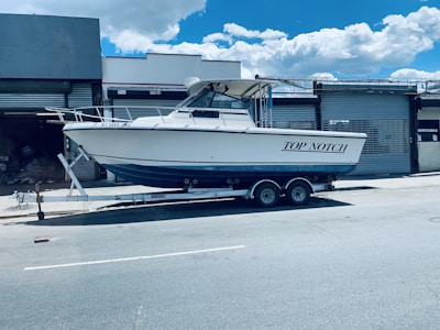 A white motorboat named 'Top Notch' is parked on a trailer in an industrial area. The trailer is positioned on a street in front of a building with metal shutters. The sky is clear with a few clouds.