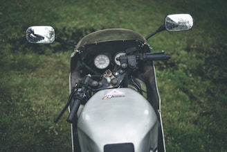 A close-up view of a motorcycle's handlebars and dashboard, set against a grassy background. The side mirrors reflect the surrounding greenery, and the instrumentation panel is clearly visible.