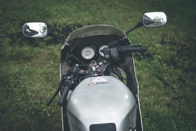 A close-up view of a motorcycle's handlebars and dashboard, set against a grassy background. The side mirrors reflect the surrounding greenery, and the instrumentation panel is clearly visible.