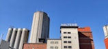 A large industrial building complex with several silos and a structure labeled 'MALTERIE'. The architecture includes a mix of gray silos and rectangular buildings in shades of red and beige against a clear blue sky.