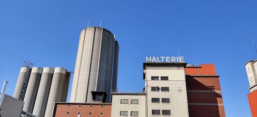 A large industrial building complex with several silos and a structure labeled 'MALTERIE'. The architecture includes a mix of gray silos and rectangular buildings in shades of red and beige against a clear blue sky.