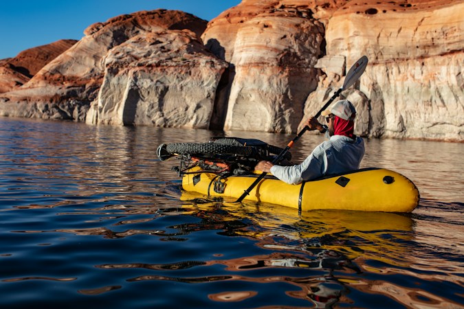 Paddler in a yellow packraft on calm blue water below sandstone cliffs, a bicycle and gear lashed across the bow