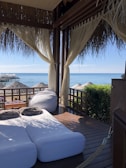 A cozy outdoor lounging area with two cushioned sunbeds and a bean bag under a thatched-roof cabana. Sheer curtains frame the view, which includes a wooden deck overlooking a beach with straw umbrellas and the calm ocean water. There is greenery to the right of the cabana and a wooden table with folded towels on it.