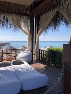 A cozy outdoor lounging area with two cushioned sunbeds and a bean bag under a thatched-roof cabana. Sheer curtains frame the view, which includes a wooden deck overlooking a beach with straw umbrellas and the calm ocean water. There is greenery to the right of the cabana and a wooden table with folded towels on it.