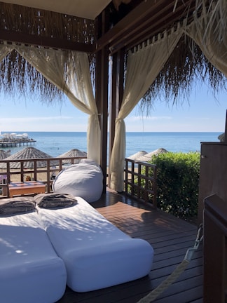 A cozy outdoor lounging area with two cushioned sunbeds and a bean bag under a thatched-roof cabana. Sheer curtains frame the view, which includes a wooden deck overlooking a beach with straw umbrellas and the calm ocean water. There is greenery to the right of the cabana and a wooden table with folded towels on it.