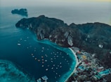 An aerial view of a coastal landscape featuring turquoise waters, a rocky island with lush greenery, and multiple boats scattered across the water. There's a small town or village with colorful buildings along the shoreline, backed by forested hills.