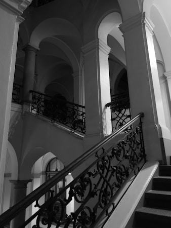 An ornate interior staircase with elegant wrought iron railings and large arched openings, creating a striking play of light and shadow in a monochrome setting.