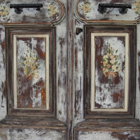 An antique wooden door with ornate floral decorations is prominently featured. The surface is weathered, showcasing a patina of faded paint and exposed wood grain. The floral motifs are delicately painted, with clusters of roses and greenery, adding a vintage charm to the doors.