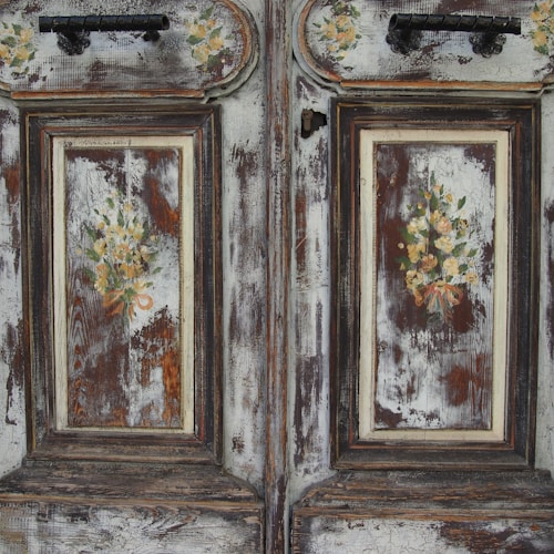 An antique wooden door with ornate floral decorations is prominently featured. The surface is weathered, showcasing a patina of faded paint and exposed wood grain. The floral motifs are delicately painted, with clusters of roses and greenery, adding a vintage charm to the doors.