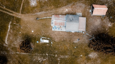An aerial view of a rural area featuring a house with a rusty, patchy roof surrounded by open grassy fields. The landscape includes a few scattered trees and another smaller building nearby. Unpaved paths and dispersed items are visible across the scene, giving a sense of isolation and simplicity.