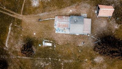 An aerial view of a rural area featuring a house with a rusty, patchy roof surrounded by open grassy fields. The landscape includes a few scattered trees and another smaller building nearby. Unpaved paths and dispersed items are visible across the scene, giving a sense of isolation and simplicity.