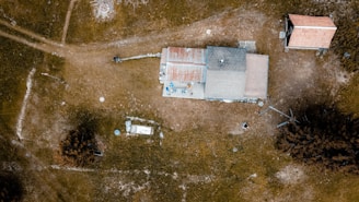 An aerial view of a rural area featuring a house with a rusty, patchy roof surrounded by open grassy fields. The landscape includes a few scattered trees and another smaller building nearby. Unpaved paths and dispersed items are visible across the scene, giving a sense of isolation and simplicity.