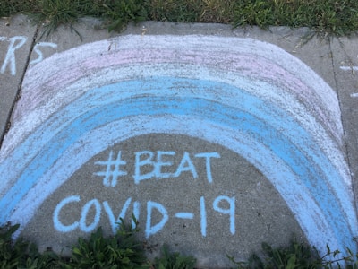 A colorful chalk drawing on a concrete sidewalk features a rainbow predominantly in shades of blue and pink. The words #BEAT COVID-19 are written in large letters underneath the rainbow. Grass is visible along the edges of the sidewalk.
