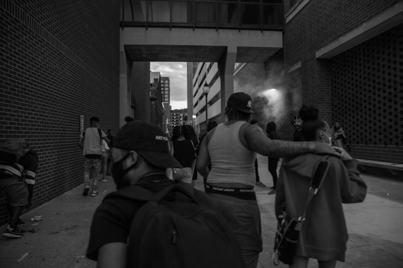 A group of people walking through an urban alleyway surrounded by tall brick buildings. Some individuals are wearing caps and one person is carrying a backpack. The scene appears to be dimly lit, with smoke or mist visible near the side of a building in the background.