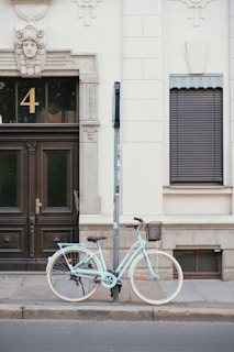 A light blue bicycle is securely locked to a pole on a sidewalk in front of a classic building facade featuring a dark wooden door with decorative molding and a number 4 above it. The wall is elegantly designed with an ornamental face at the top and a window with closed shutters.