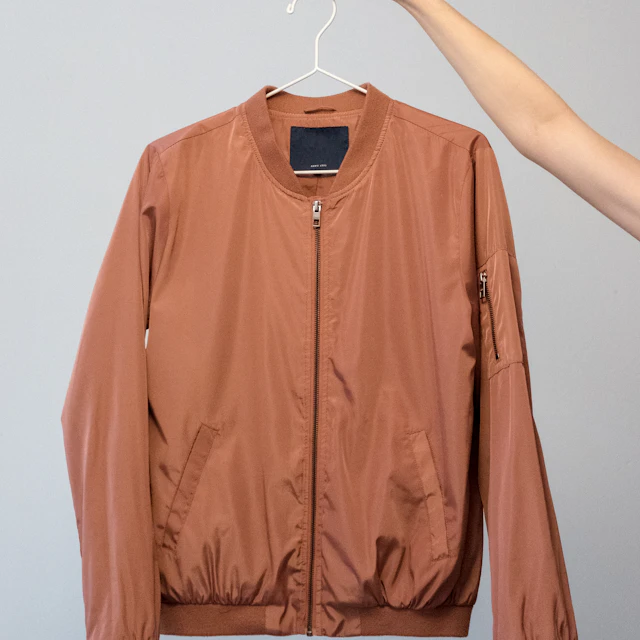 A hand holding a brown bomber jacket on a white hanger against a wall