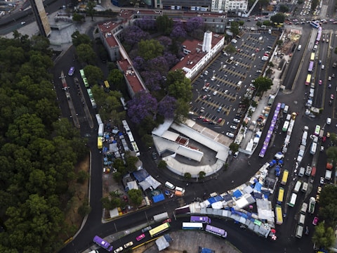 An aerial view of a bustling urban area featuring a large parking lot with several vehicles, a central building with a tall white structure, and surrounding trees with purple foliage. A busy street with numerous buses and cars runs adjacent to the parking area. Temporary structures or market stalls are set up along the street near the buses.