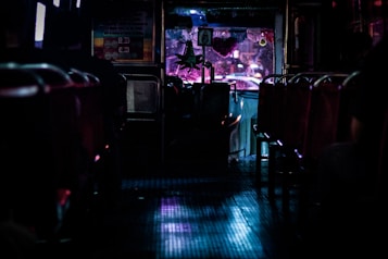 A dimly lit interior of a bus with empty seats, leading to the driver's area. The lights from outside cast a purple and blue glow onto the floor. Decorations hang near the front window, and blurred city lights are visible through the windshield.