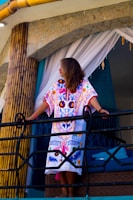 A woman standing on a balcony, wearing a colorful embroidered dress with intricate patterns. She is leaning on a black railing, surrounded by bamboo poles and stone walls, with white and teal curtains in the background.