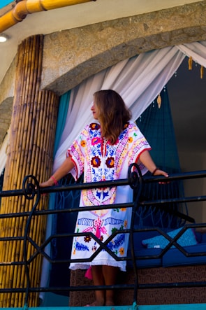 A woman standing on a balcony, wearing a colorful embroidered dress with intricate patterns. She is leaning on a black railing, surrounded by bamboo poles and stone walls, with white and teal curtains in the background.