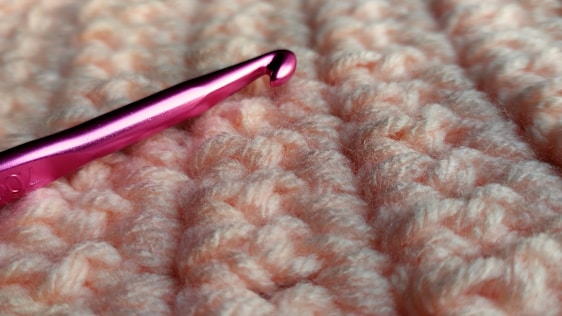A close-up view of a pink crochet hook resting on a textured, peach-colored yarn fabric. The hook has a metallic sheen and is prominently positioned against the soft, knotted surface of the fabric.