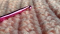 A close-up view of a pink crochet hook resting on a textured, peach-colored yarn fabric. The hook has a metallic sheen and is prominently positioned against the soft, knotted surface of the fabric.