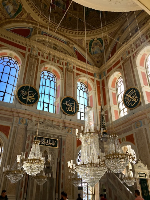 An ornate interior with high, vaulted ceilings and large arched windows. Crystal chandeliers hang from the ceiling, illuminating the space. Decorative Islamic calligraphy is prominently displayed on circular plaques. The walls and ceiling feature intricate patterns and artwork.