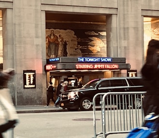 A city scene featuring the entrance of a television studio. The signage for 'The Tonight Show Starring Jimmy Fallon' is illuminated in bright colors above the entrance. A vehicle is parked in front, and several pedestrians are visible on the sidewalk. The building facade displays detailed art deco murals.