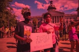 A group of people participating in a protest in front of a large government building with columns and a dome. Two individuals in the foreground hold a sign advocating for justice and the Black Lives Matter movement. The crowd is diverse, with some participants wearing masks and holding up their fists in solidarity.