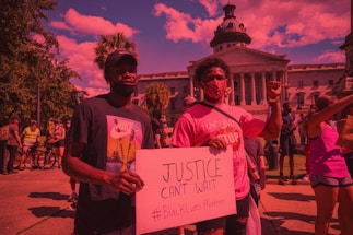 A group of people participating in a protest in front of a large government building with columns and a dome. Two individuals in the foreground hold a sign advocating for justice and the Black Lives Matter movement. The crowd is diverse, with some participants wearing masks and holding up their fists in solidarity.