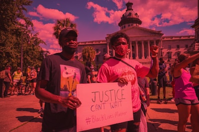 A group of people participating in a protest in front of a large government building with columns and a dome. Two individuals in the foreground hold a sign advocating for justice and the Black Lives Matter movement. The crowd is diverse, with some participants wearing masks and holding up their fists in solidarity.