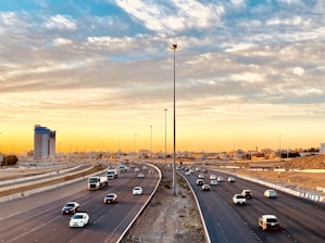 An expansive highway with multiple lanes lined with vehicles traveling in both directions. The highway is flanked by a construction site on one side and a few buildings, including a prominent skyscraper, on the other. The sky is filled with scattered clouds, illuminated by the warm colors of either sunrise or sunset, casting a golden hue over the scene.