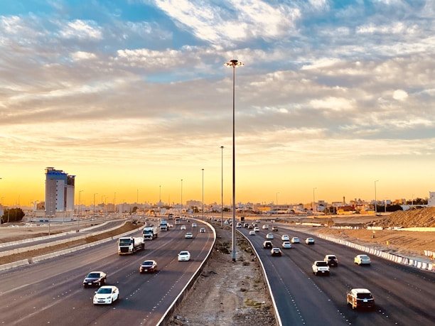 An expansive highway with multiple lanes lined with vehicles traveling in both directions. The highway is flanked by a construction site on one side and a few buildings, including a prominent skyscraper, on the other. The sky is filled with scattered clouds, illuminated by the warm colors of either sunrise or sunset, casting a golden hue over the scene.