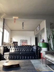 A modern living room with a black leather tufted sofa as the central piece. On the left, a row of windows lets in natural light. There are decorative elements like a white brick wall with abstract art, a wooden coffee table slab, and a potted cactus plant. The room also features a metallic table with bottles in the corner and a grey upholstered chair on the right.