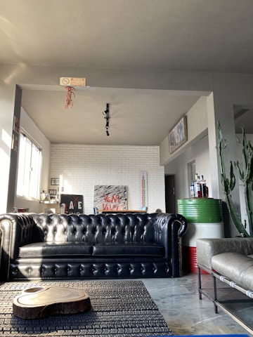 A modern living room with a black leather tufted sofa as the central piece. On the left, a row of windows lets in natural light. There are decorative elements like a white brick wall with abstract art, a wooden coffee table slab, and a potted cactus plant. The room also features a metallic table with bottles in the corner and a grey upholstered chair on the right.